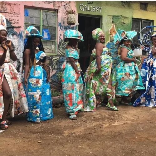 Women in vibrant, colorful dresses made of recycled materials stand joyfully in front of an art gallery.