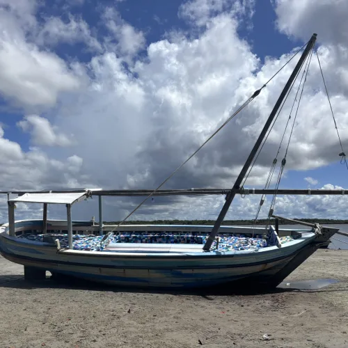Traditional wooden dhow boat resting on sandy shore with ocean and cloudy sky in Kenya - Flipflopi Dhow